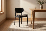 A black Simone dining chair on a woven wool rug beside a light oak dining table with a ribbed glass and an olive branch in a stoneware vase, in warm sunlight from a window