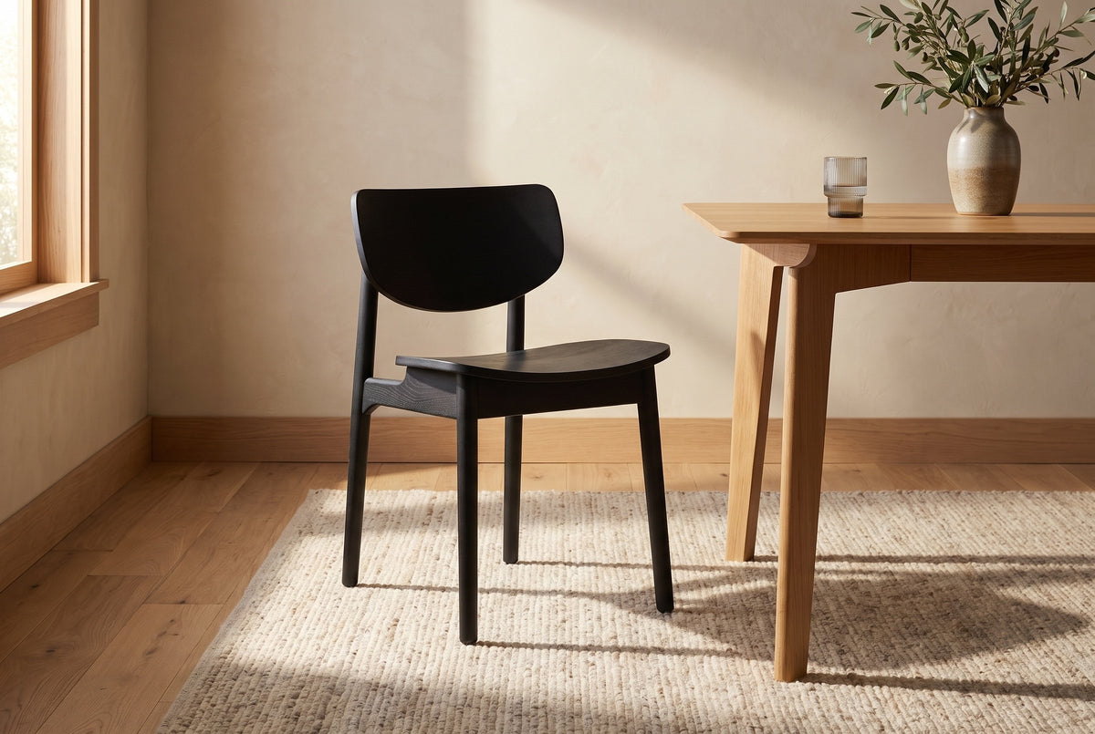 A black Simone dining chair on a woven wool rug beside a light oak dining table with a ribbed glass and an olive branch in a stoneware vase, in warm sunlight from a window