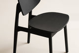 Close-up of a black Simone dining chair on a warm off-white studio backdrop: the curved back panel on a round rear post, the contoured oak seat and the notched side rail, in matte black with fine oak grain