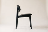 Black Simone dining chair in side view on a plain warm off-white studio backdrop, showing the curved back panel raised on the rear posts, the open gap above the seat, the side rail and four slim legs