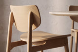 Close-up of a natural oak Simone chair from behind: the curved back panel on two round posts and the oak grain across the seat, beside a travertine table