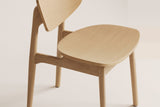 Natural honey oak Simone chair, close-up of the contoured seat, back panel, post and side-rail joint on a plain warm off-white studio backdrop