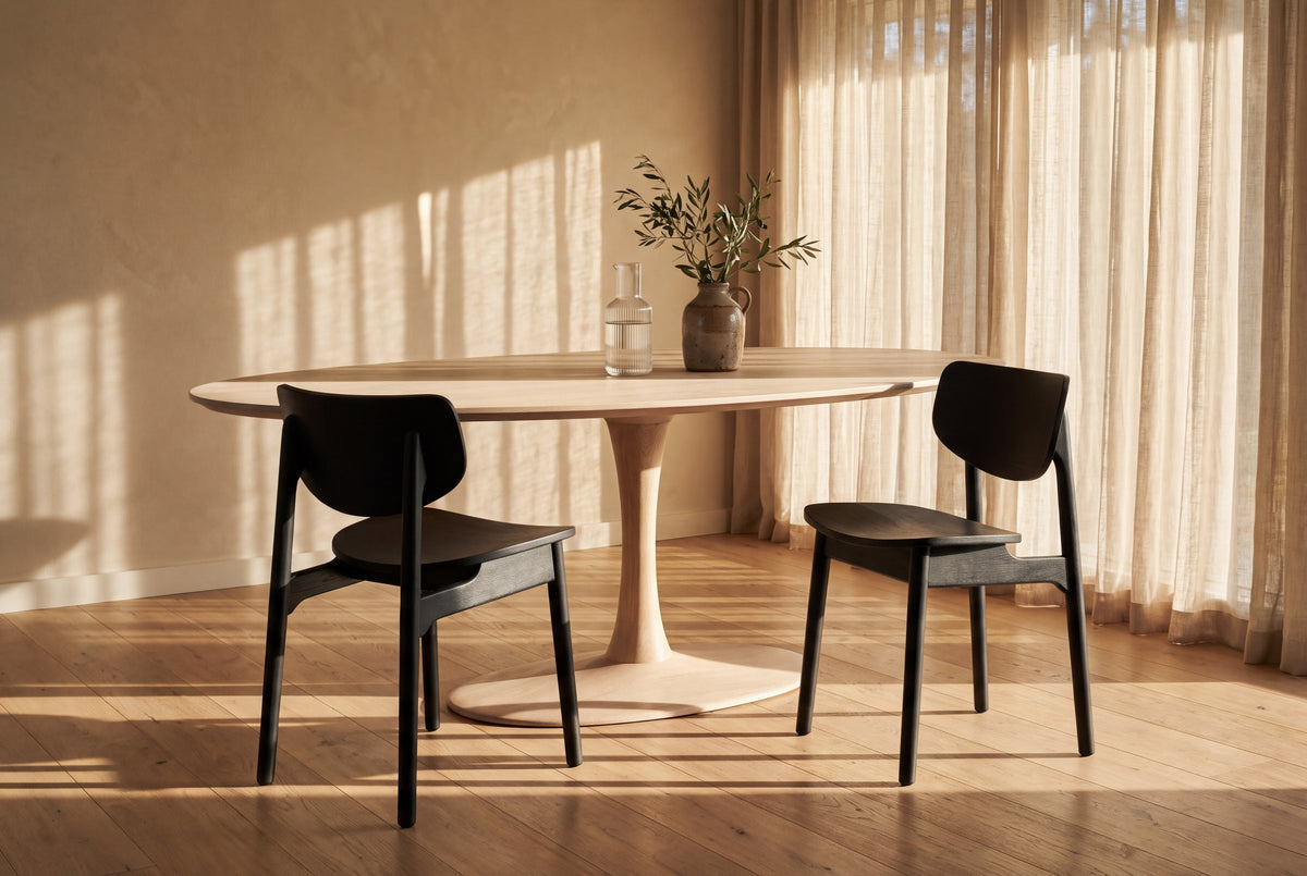 Two black Simone dining chairs at an oval white oak pedestal table with a glass carafe and an olive branch in a stoneware jug, in a dining room with sheer linen curtains, a warm plaster wall and low sun across a wide-plank oak floor