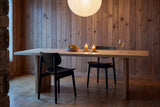 Two black Simone dining chairs at a long oak table with a bowl of pears, two lit candles and a glass of white wine, under a paper lantern against a knotty wood-panelled wall in a cottage at dusk, with a fieldstone wall at the edge