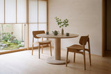 Two natural oak Simone dining chairs at a round pale wood pedestal table with a celadon tea set and a maple branch, in a Kyoto room with paper screens and a moss garden