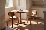 Two natural oak Simone dining chairs at a round oak pedestal table with a bowl of lemons, a glass of water and a linen napkin, in a sunlit breakfast nook with white plaster walls, an arched window and pale kitchen cabinets