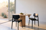 Two black Simone dining chairs at a round travertine pedestal table with a bowl of limes, a glass carafe and a linen napkin, on a terrazzo floor beside sliding glass doors onto a sunlit courtyard with an olive tree and agaves