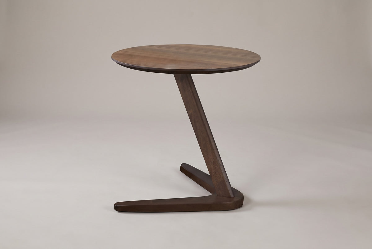 Vigevano C-shaped side table in walnut on a warm grey studio backdrop: a round top on one leaning post over a boomerang base
