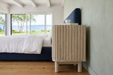 Side view of the Vitorchiano nightstand in Natural White Wash in a Metis-sur-Mer summer bedroom, with sage limewash walls