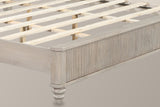 Close-up of the beige reeded footboard corner with its rounded cap and a turned bun foot, the pale slats behind